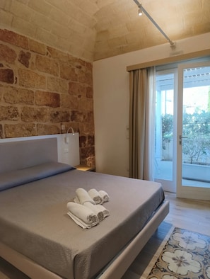 A bedroom with a stone wall, a bed with a grey mattress, and a view of the outdoors through a sliding door. - Butiko (Castelluzzo)