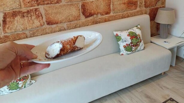 Eine Hand hält einen Teller mit Dessert, ein weißes Sofa und eine Backsteinwand im Hintergrund.
