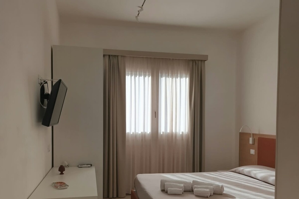 A hotel room with a bed, a desk, a mirror, and a window with curtains.
