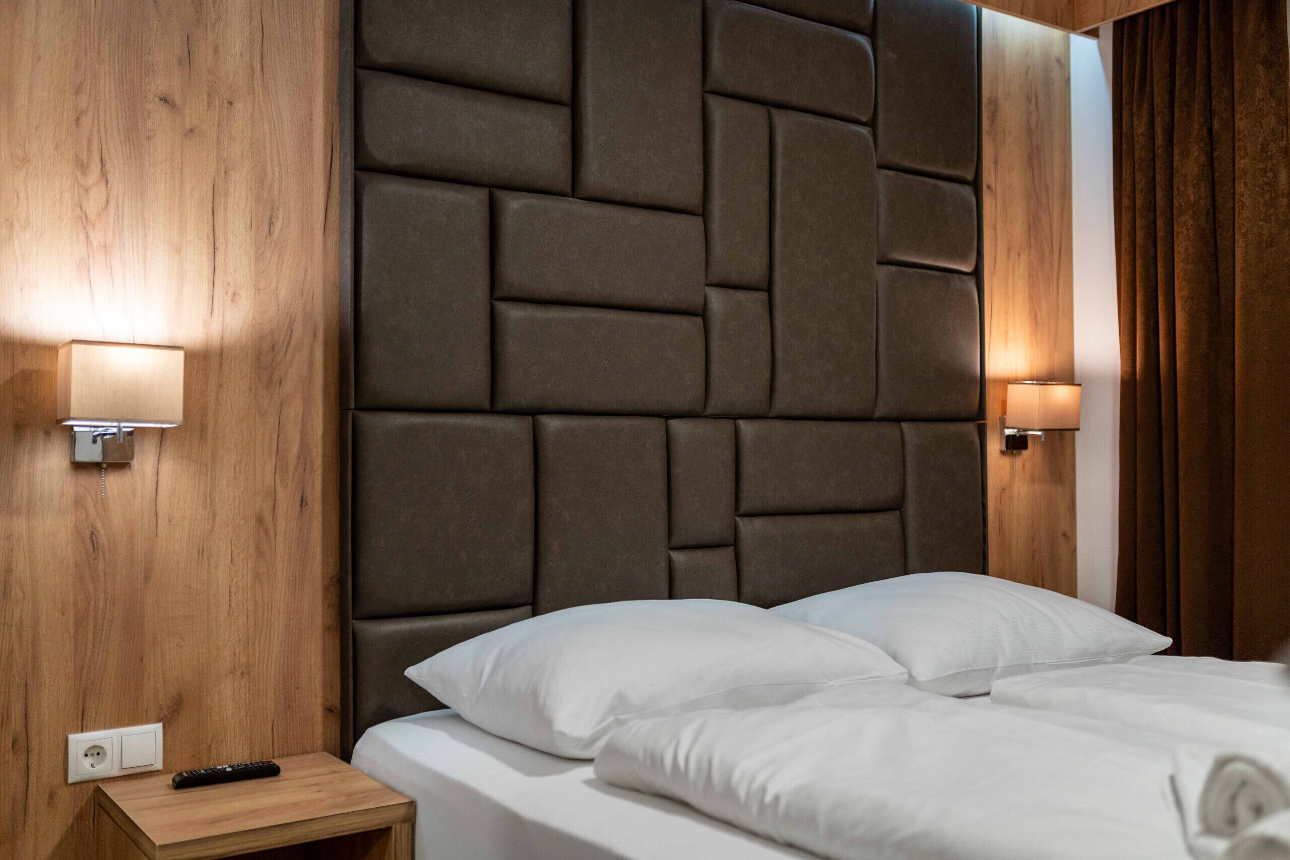 Une chambre d’hôtel avec une tête de lit en bois, deux lampes de chevet, une table de chevet avec une télécommande et un lit parfaitement fait.