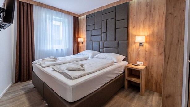 A hotel room with a bed, wooden headboard, two bedside lamps, a TV, and a small wooden nightstand. - Maiskogelblick Apartments - TOP 3, Garden & Sauna (Piesendorf)