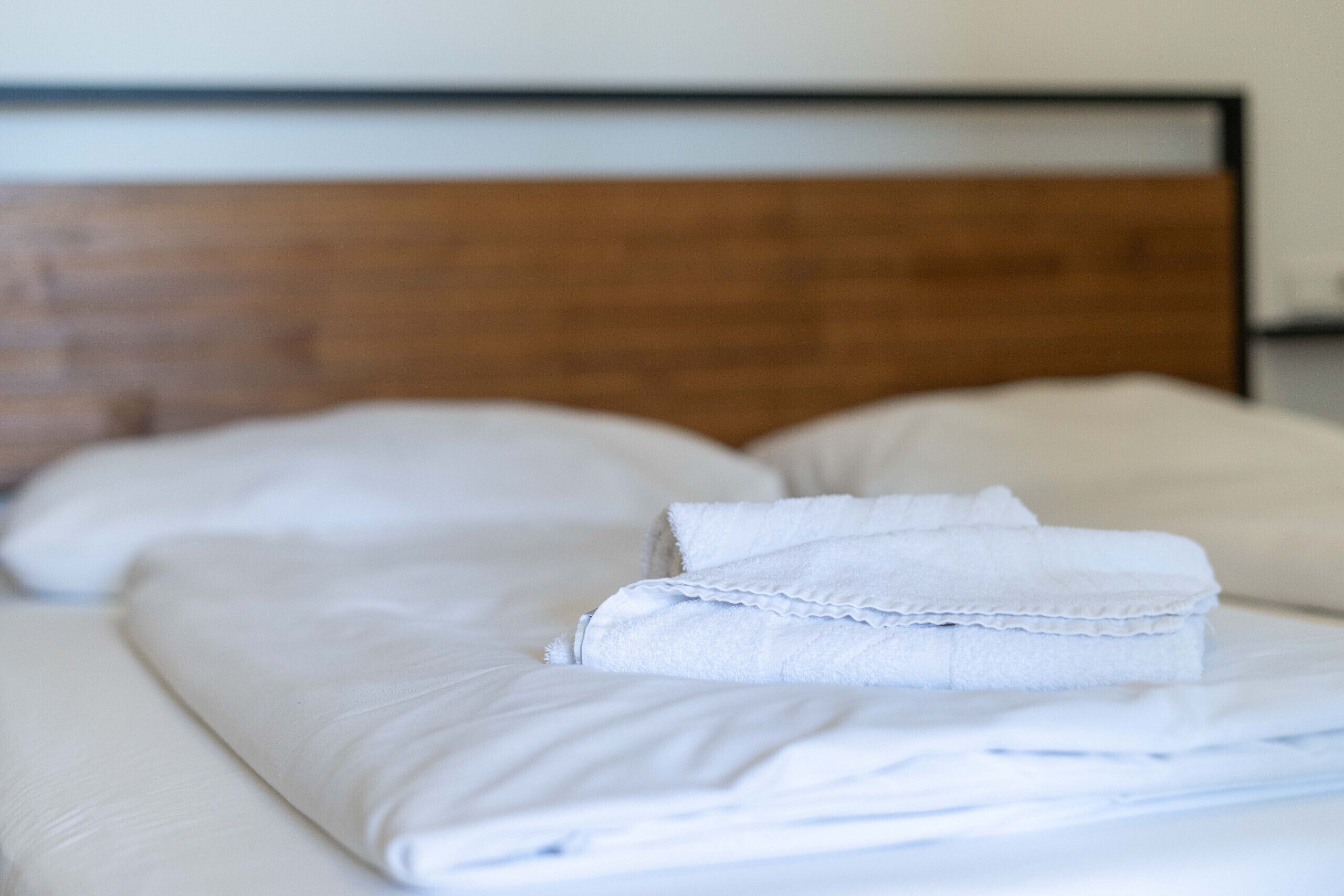 Ein ordentlich bezogenes Bett mit weißen Leinen und Kissen, ein hölzerner Kopfteil und ein schlichtes, aufgeräumtes Zimmerdesign.