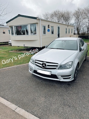 Um Mercedes-Benz branco estacionado em frente a uma casa móvel com uma placa que diz 'Gary's paradise'.