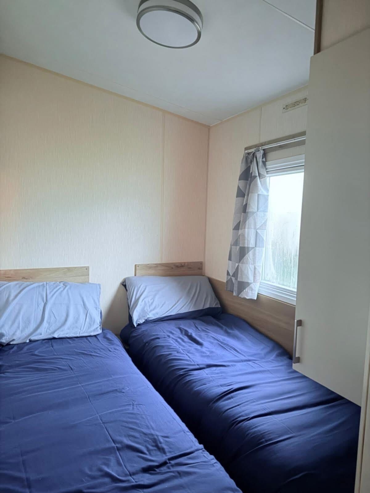 A room with two beds, a window with a curtain, and a ceiling light fixture.
