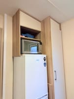 Una kitchenette compacta con un microondas sobre un refrigerador blanco, armarios de madera y un estante pequeño.