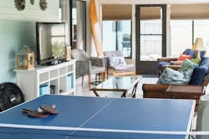 A ping pong table in a living room with a TV, chairs, and a surfboard. - Sea Breeze Sanctuary (Ormond Beach)