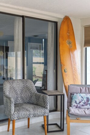 A room with a patterned armchair, a small table, and a surfboard leaning against the wall. - Sea Breeze Sanctuary (Ormond Beach)