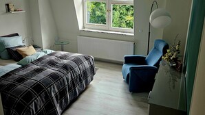 Un dormitorio con cama, una silla, una mesa, un ventanal con vistas a zonas verdes y un radiador.