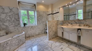A bathroom with marble walls, a bathtub, a sink with a mirror, and a window with a view of greenery.