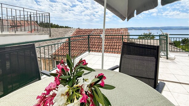 En terrass med ett bord, stolar, en blomsteruppsättning och utsikt över havet och byggnaderna.
