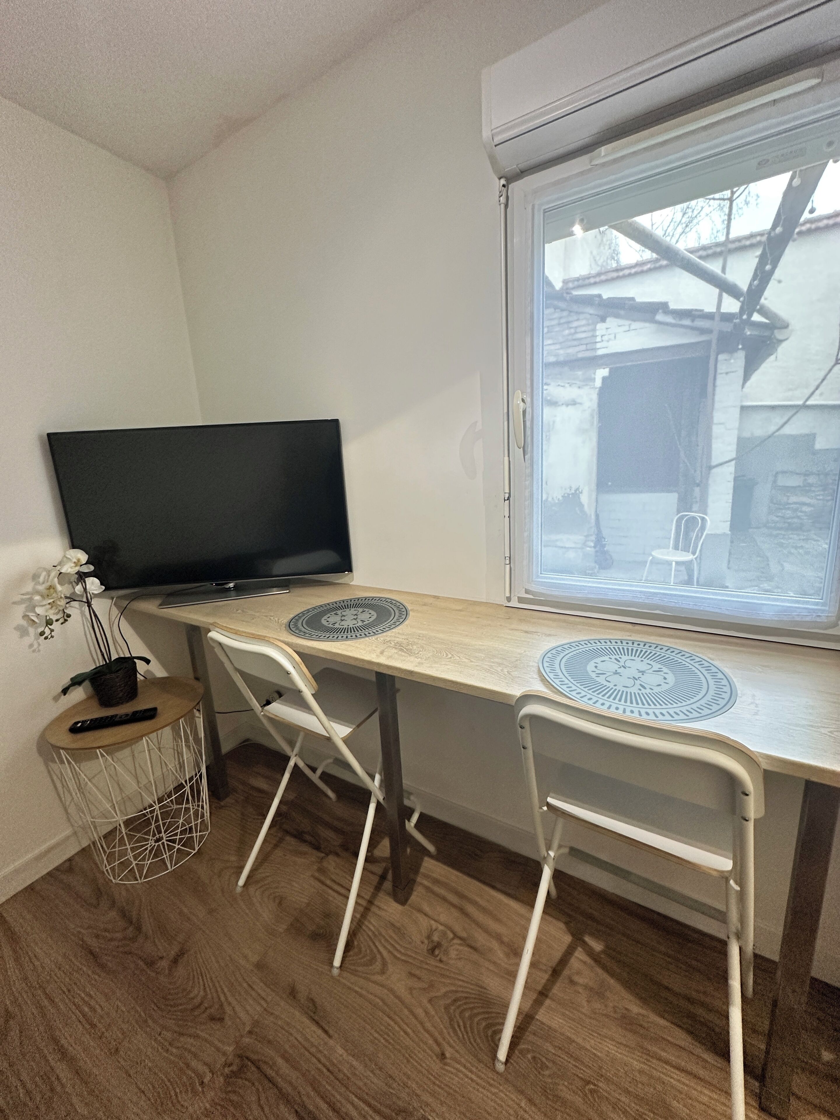 Een kamer met een houten bureau, twee witte stoelen, een televisie, een raam en een kleine plant.