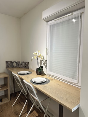 Un coin repas avec une table en bois, des chaises blanches, un panneau décoratif portant l’inscription « HOME », une plante en pot et une fenêtre avec des stores.