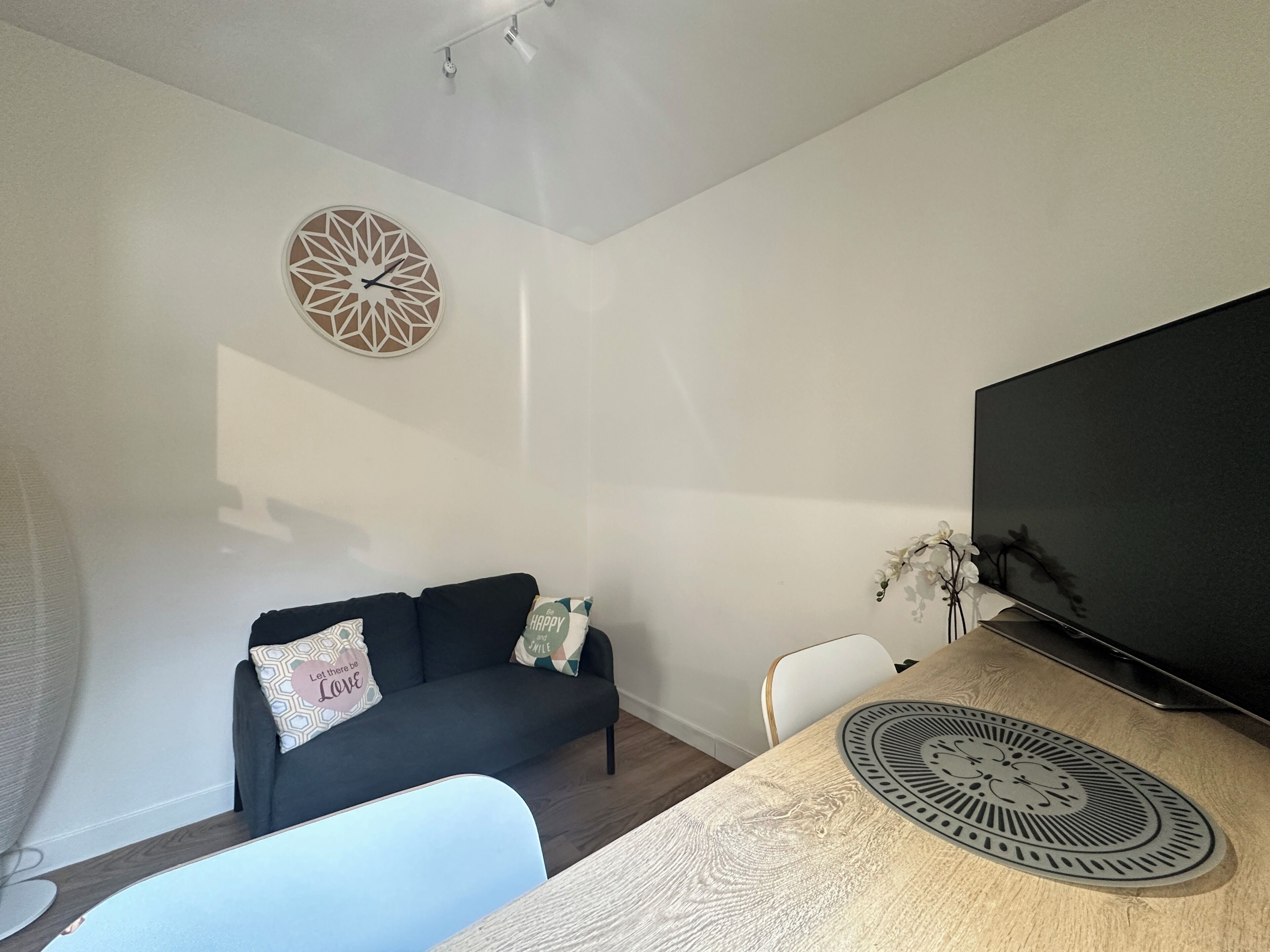 Een moderne woonkamer met een donkere bank, een ronde wandklok, een flatscreen televisie en een houten eettafel met een decoratief bord.