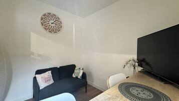 Een moderne woonkamer met een donkere bank, een ronde wandklok, een flatscreen televisie en een houten eettafel met een decoratief bord.