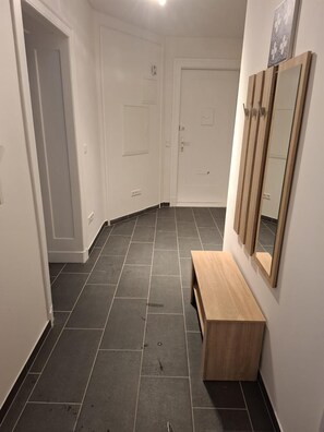 Un pasillo con suelo de baldosas oscuras, paredes blancas y un banco de madera.
