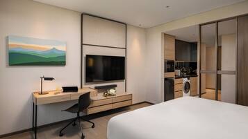 A modern hotel room with a bed, a desk with a computer, a TV, a washing machine, and a painting on the wall.