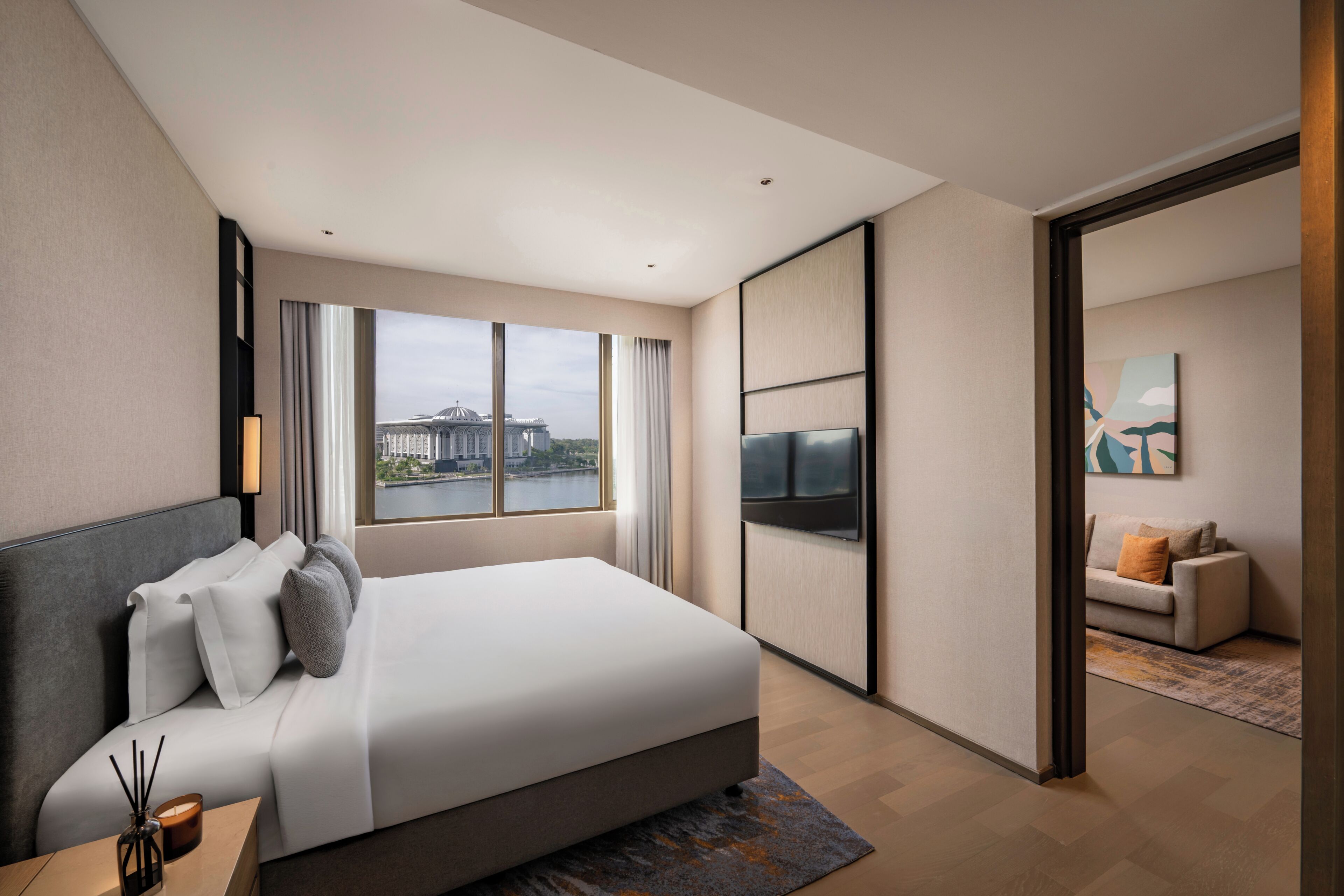 Une chambre d’hôtel moderne avec un grand lit, une télévision, une vue sur un immeuble par la fenêtre et un canapé dans la pièce adjacente.