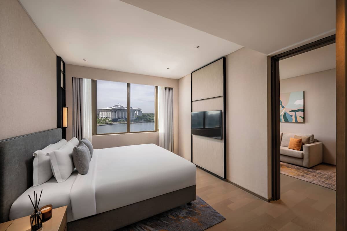 A modern hotel room with a large bed, a TV, a view of a building through the window, and a sofa in the adjacent room.