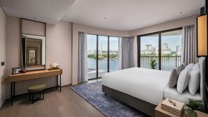 A modern hotel room with a large bed, a desk, a chair, a large window with a view of a body of water and buildings, and a balcony.
