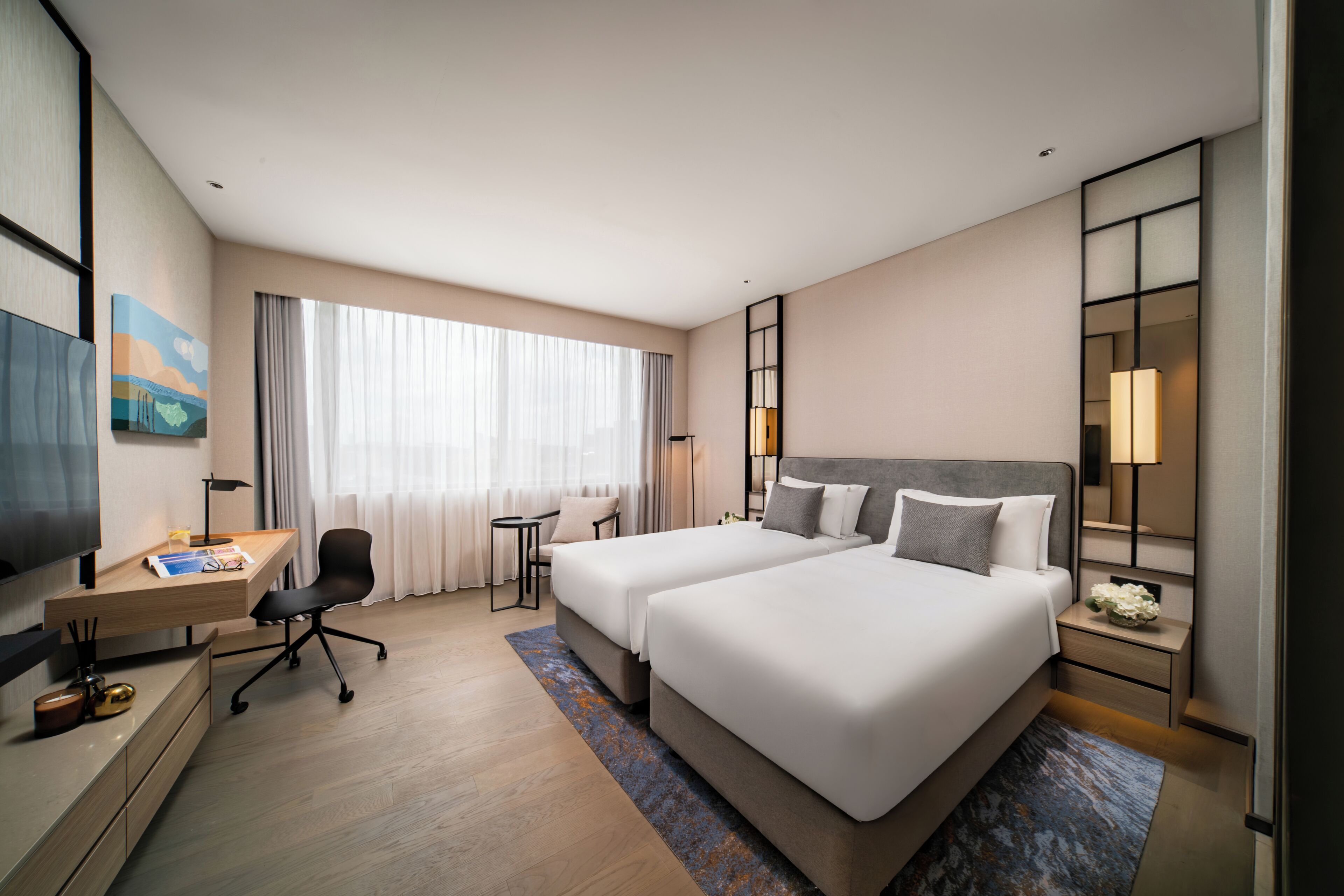 Ein Hotelzimmer mit zwei Betten, einem Schreibtisch, einem Stuhl, einem Fernseher und einem großen Fenster mit Vorhängen.