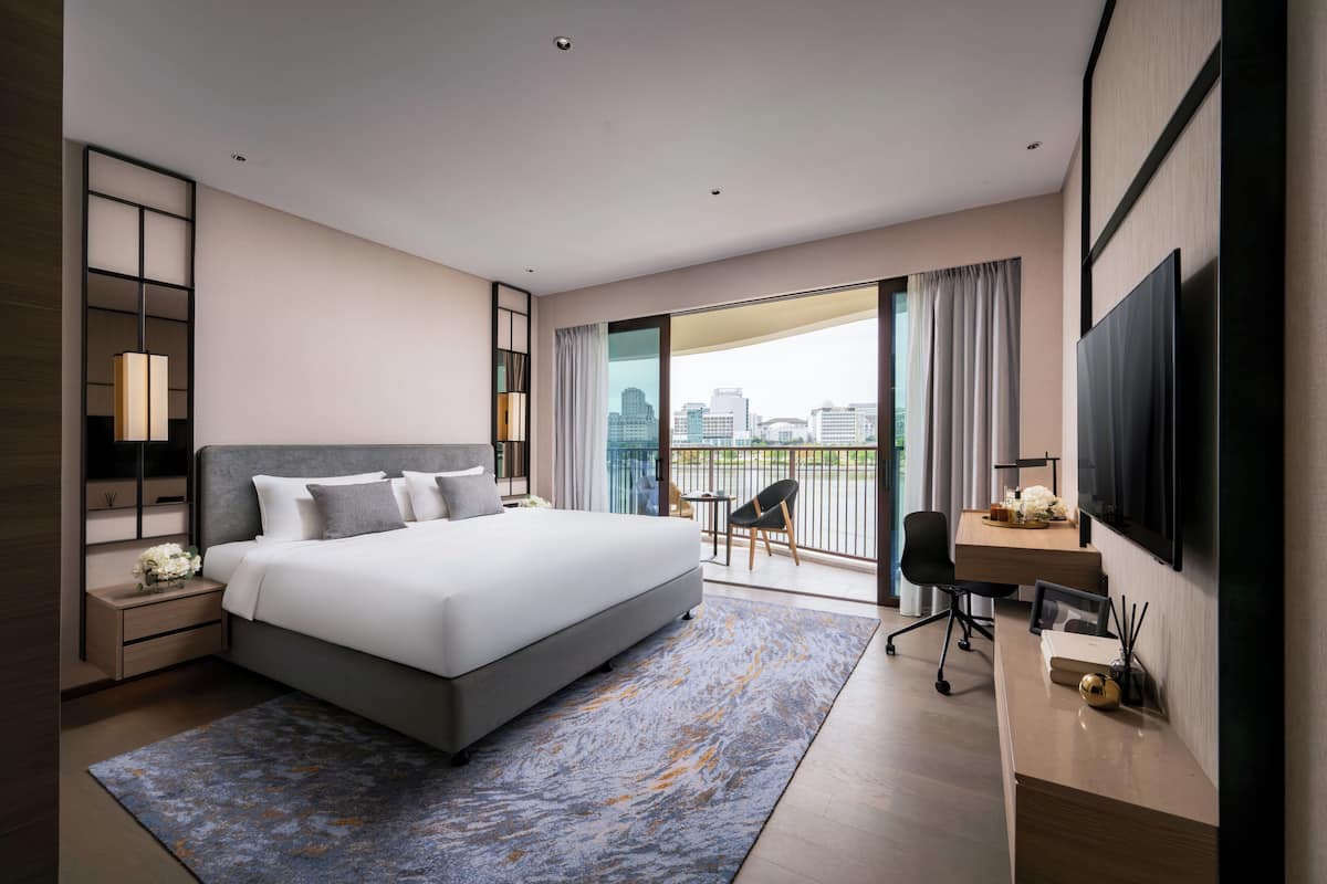 A modern hotel room with a large bed, a desk, a TV, and a balcony with a city view.