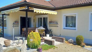 Ein Haus mit überdachter Terrasse, Gartenmöbeln, Topfpflanzen und einem kleinen Garten.