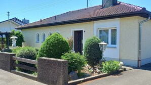 Ein eingeschossiges Gebäude mit einem Ziegeldach, einer Haustür und einem kleinen Garten mit Holzzaun und verschiedenen Pflanzen.