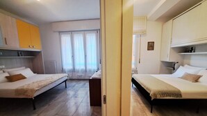 Camera d'albergo con due letti, pavimento in legno, vista attraverso una porta e una finestra con tende.
