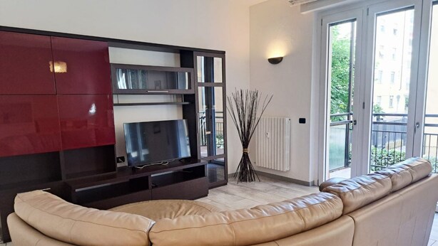 Un soggiorno con un ampio divano beige, una televisione a schermo piatto su un mobile in legno scuro, un termosifone e vista su un balcone con piante.