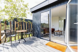 Um pátio moderno ao ar livre com deck de madeira, cadeiras de metal e porta de vidro que dá para uma área de refeições interna.