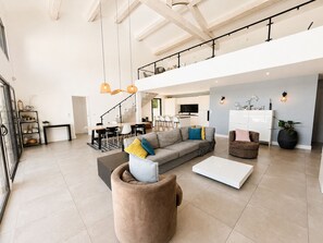 Un grand espace de vie comprenant un canapé, un pouf et une table basse, un escalier et un coin cuisine.