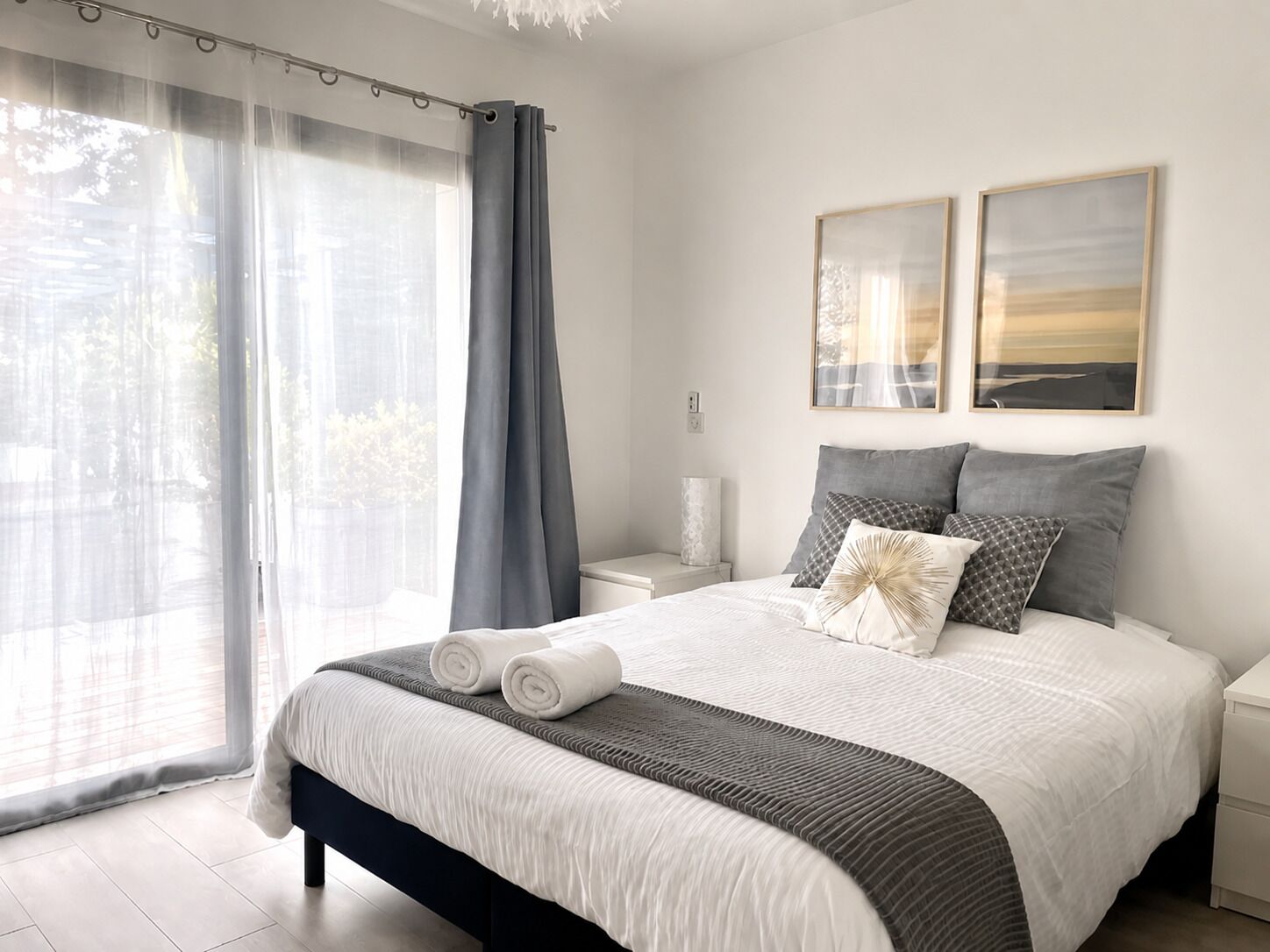 Ein ordentlich bezogenes Bett mit Kissen und Decke, ein Nachttisch mit Lampe und ein Fenster mit Vorhängen.