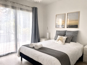 A neatly made bed with pillows and a blanket, a bedside table with a lamp, and a window with curtains.