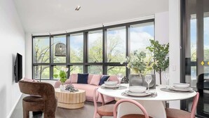 A modern living room with a sofa, a dining table set for four, and a large window with a view of trees. - LaneStay - 2BR Albert Park Views, Pool Gym Parking (Melbourne)