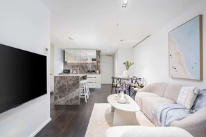 A modern living room with a flat-screen TV, a sofa, a coffee table, and a dining area with a marble island. - LaneStay - Lakeside Luxury with Pool, Gym, Parking (Melbourne)