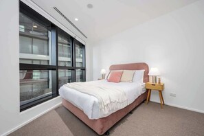 A bedroom with a large window, a bed with a pink headboard, two bedside tables with lamps, and a carpeted floor. - LaneStay - Lakeside Luxury with Pool, Gym, Parking (Melbourne)