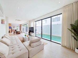 A modern living room with a large sofa, a flat-screen TV, and a view of a swimming pool through large glass doors. - Seaview Pool Villa Jomtien KTV BBQ Beach (Muang Pattaya)