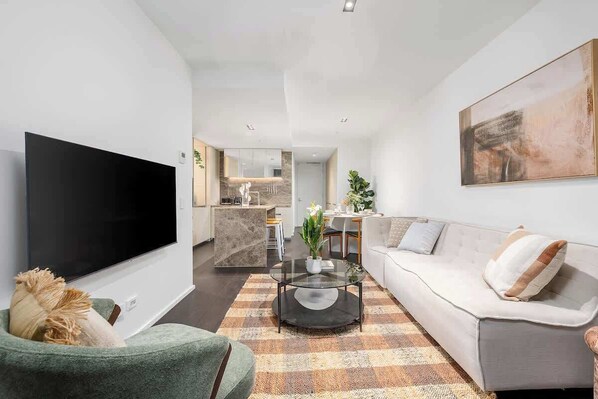 A modern living room with a flat-screen TV, a sofa, a coffee table, and a kitchen in the background. - LaneStay - Luxury Living with Pool, Gym & Parking (Melbourne)