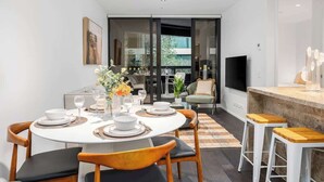 A modern dining area with a round table set for four, a bar area with stools, and a view through a glass door to an outdoor space. - LaneStay - Luxury Living with Pool, Gym & Parking (Melbourne)