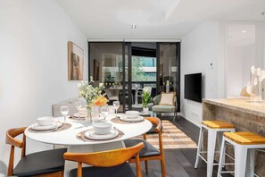 A modern dining area with a round table set for four, a bar area with stools, and a view through a glass door to an outdoor space. - LaneStay - Luxury Living with Pool, Gym & Parking (Melbourne)