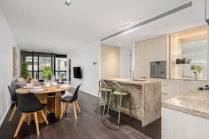 A modern kitchen with a central island, dark wooden flooring, and a dining area with a round table and chairs. - LaneStay - Lakeside Luxury w Pool, Gym & Parking (Melbourne)