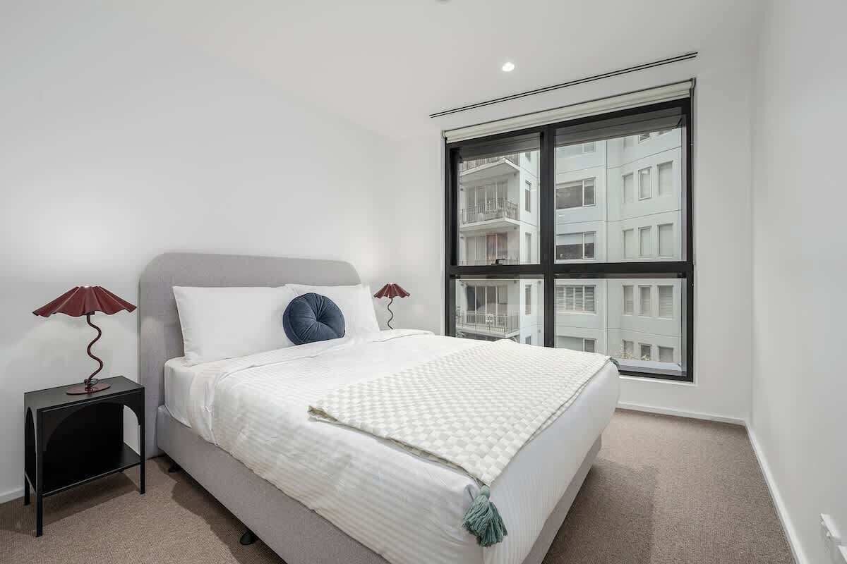 A bedroom with a bed, two bedside lamps, a window, and a view of buildings.
