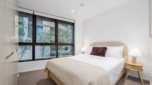 Ein Hotelzimmer mit großem Fenster, einem Bett mit weißer Bettwäsche, einem Nachttisch aus Holz und einer Lampe.