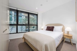 Ein Hotelzimmer mit großem Fenster, einem Bett mit weißer Bettwäsche, einem Nachttisch aus Holz und einer Lampe.
