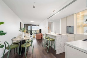 A modern kitchen with a marble countertop, a dining area with green chairs, and a large window. - LaneStay - Albert Park View w Pool, Gym & Parking (Melbourne)