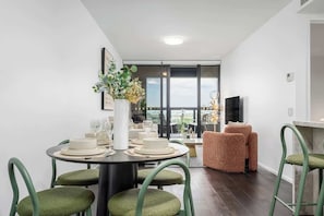 A dining area with a round table, green upholstered chairs, and a vase with flowers. - LaneStay - Albert Park View w Pool, Gym & Parking (Melbourne)