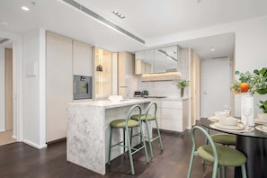 A modern kitchen with a marble island, green bar stools, and a dining area with a table set for four. - LaneStay - Albert Park View w Pool, Gym & Parking (Melbourne)