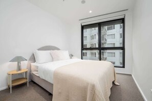 A bedroom with a bed, a nightstand, a lamp, a window with a view of buildings, and a carpeted floor. - LaneStay - Elevated 2B2B w Pool, Gym and Parking (Melbourne)