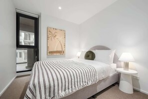 Une chambre à coucher comprenant un lit, une table de chevet, une lampe, une fenêtre et une œuvre d’art murale.