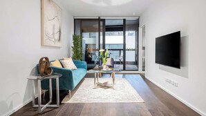 Una sala moderna con un sofá azul, una mesa de centro de cristal, un televisor de pantalla plana y un ventanal corredero que da a un balcón.